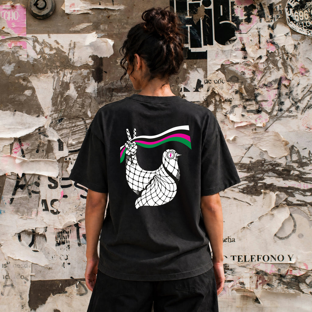 Woman wearing faded black Dove Palestinian T-shirt from the back with large keffiyeh dove artwork against a weathered poster wall.