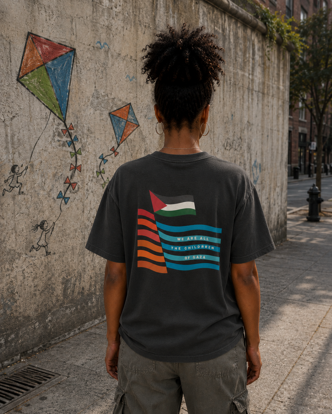 Back view of faded black heavyweight Children of Gaza Palestinian T-shirt worn outdoors beside a wall with kite mural artwork.