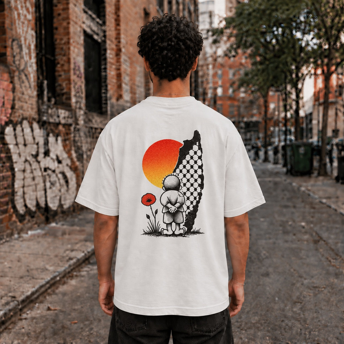 Man wearing a Faded Bone heavyweight Handala Palestinian shirt with Naji al-Ali’s Handala artwork printed on the back, standing in a city alley.