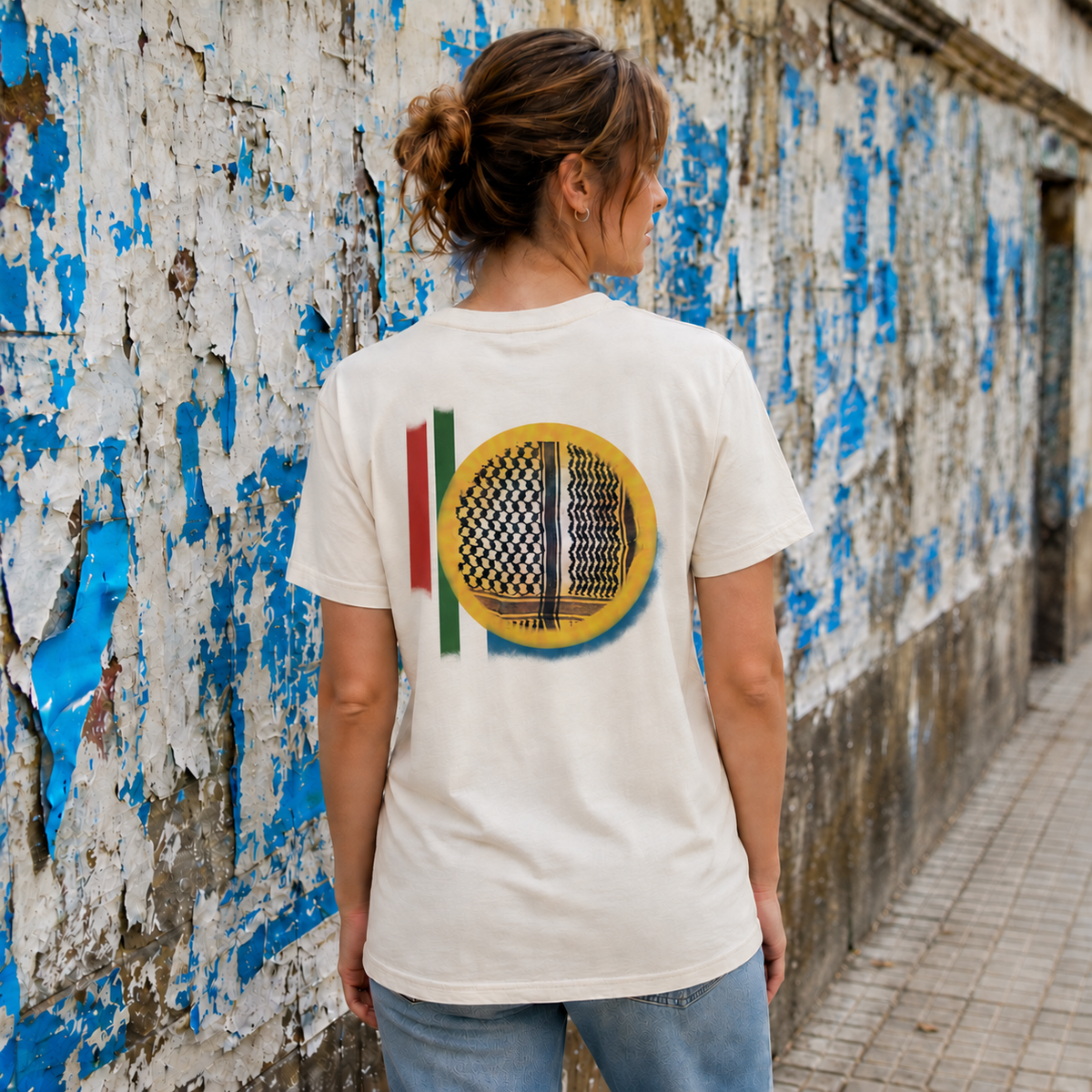 Back view of woman wearing natural Keffiyeh Sun Palestinian T-shirt against a weathered blue and white poster wall.