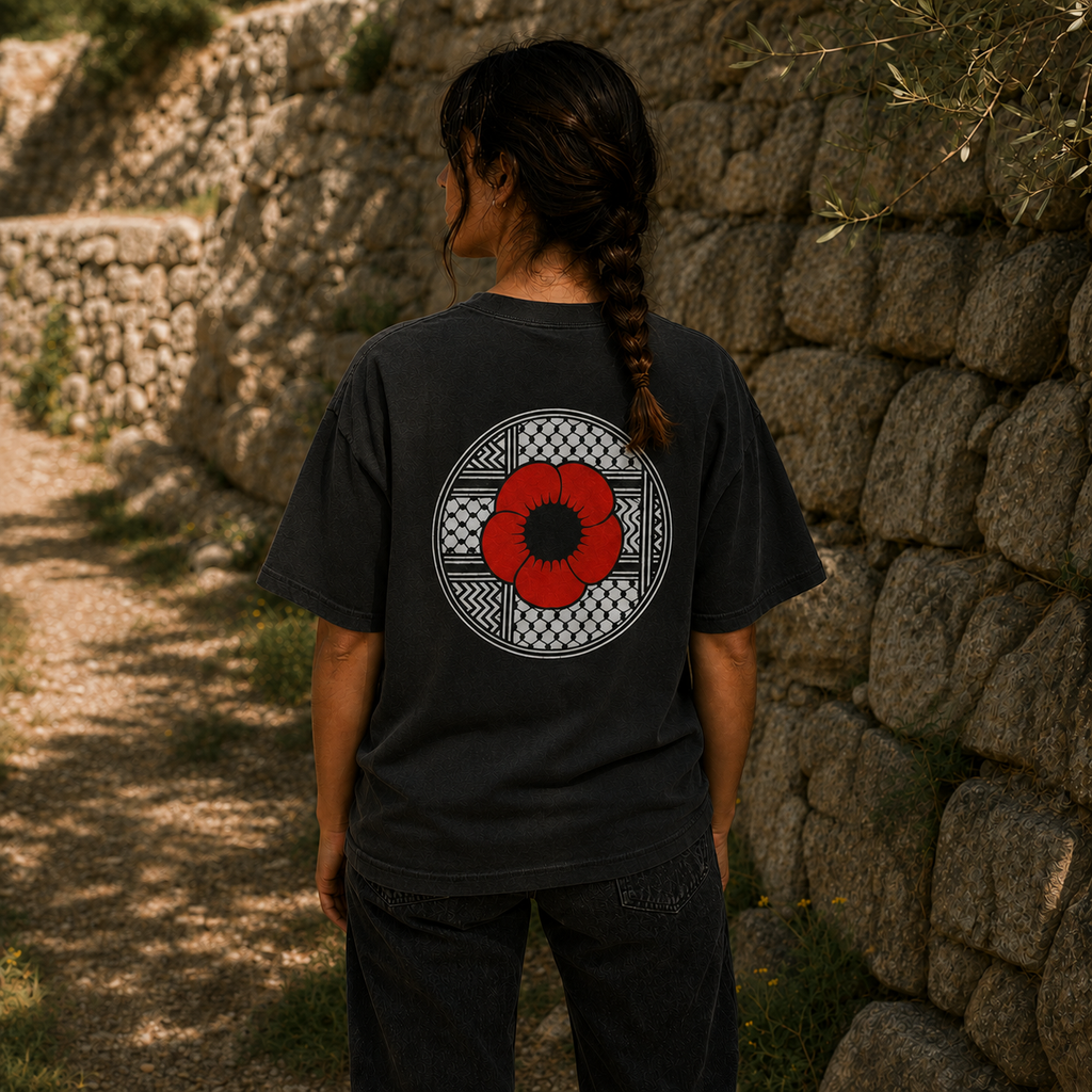 Back view of faded black heavyweight Palestinian Poppy T-shirt featuring red poppy and keffiyeh artwork, worn outdoors beside a stone wall.