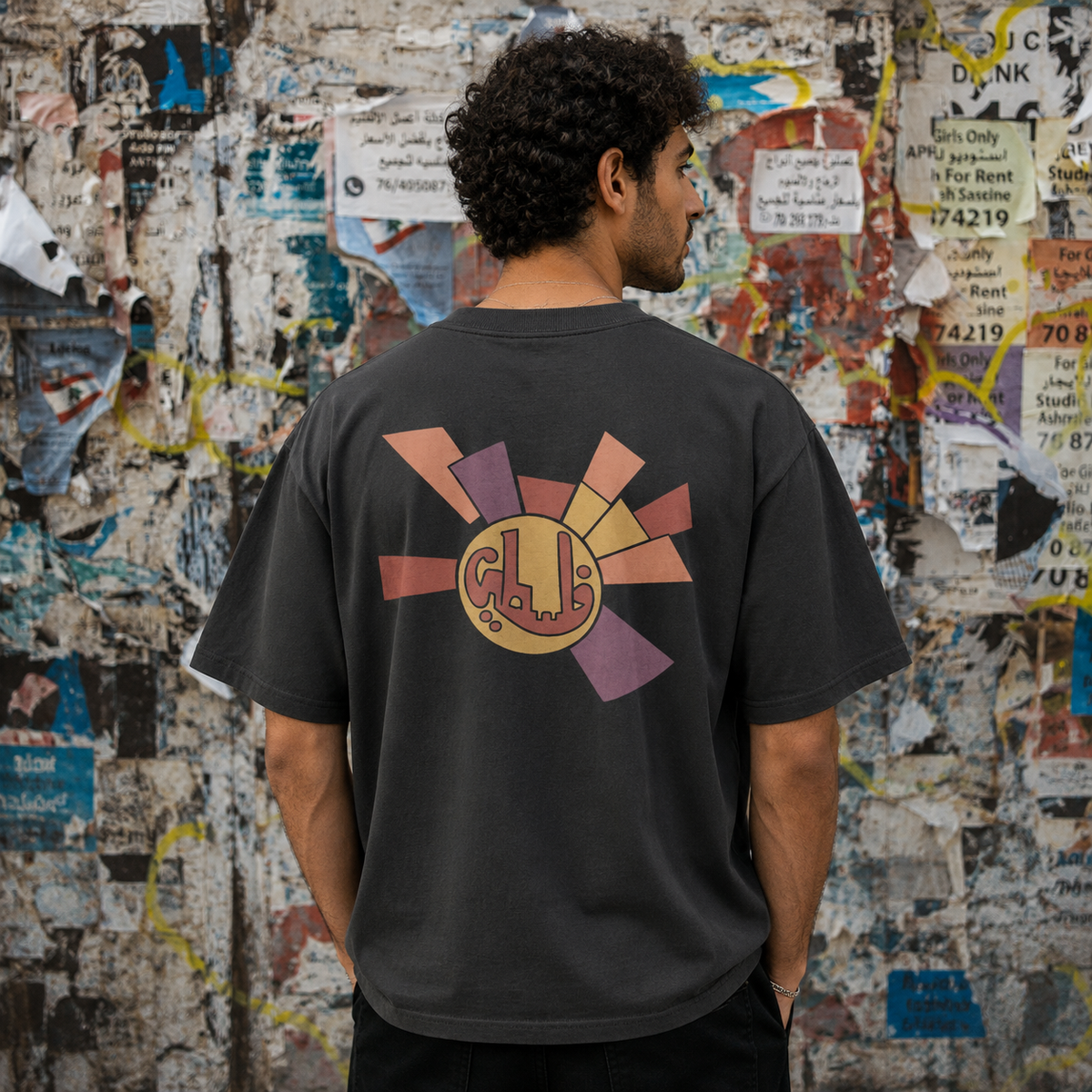 Back view of male model wearing the faded black heavyweight Sun Emblem Palestinian T-shirt in front of a distressed poster wall.