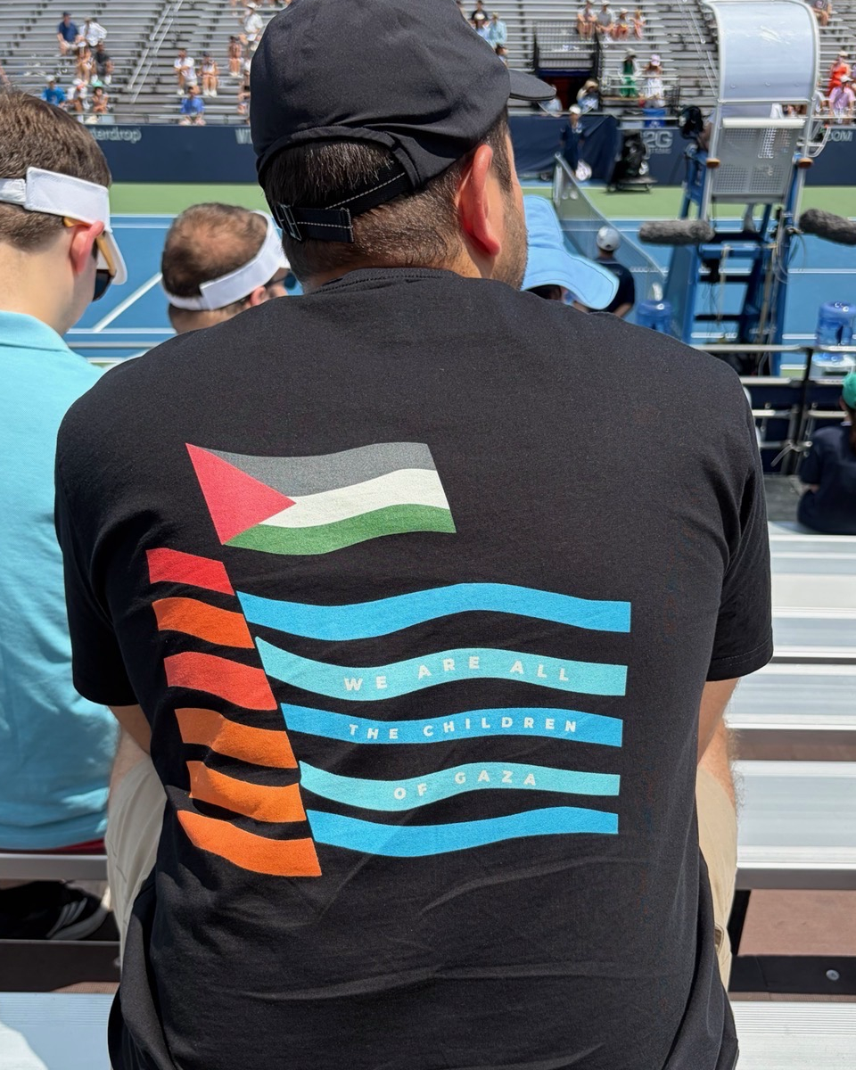 Man wearing the black heavyweight Children of Gaza Palestinian T-shirt, shown from the back while seated at a tennis match.