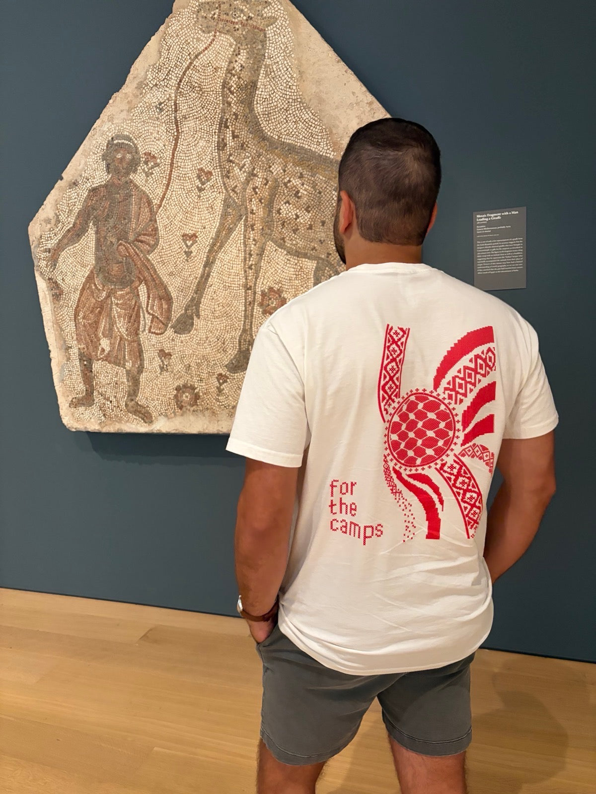 Man wearing the cream For the Camps Palestinian T-shirt, shown from the back in an art museum.