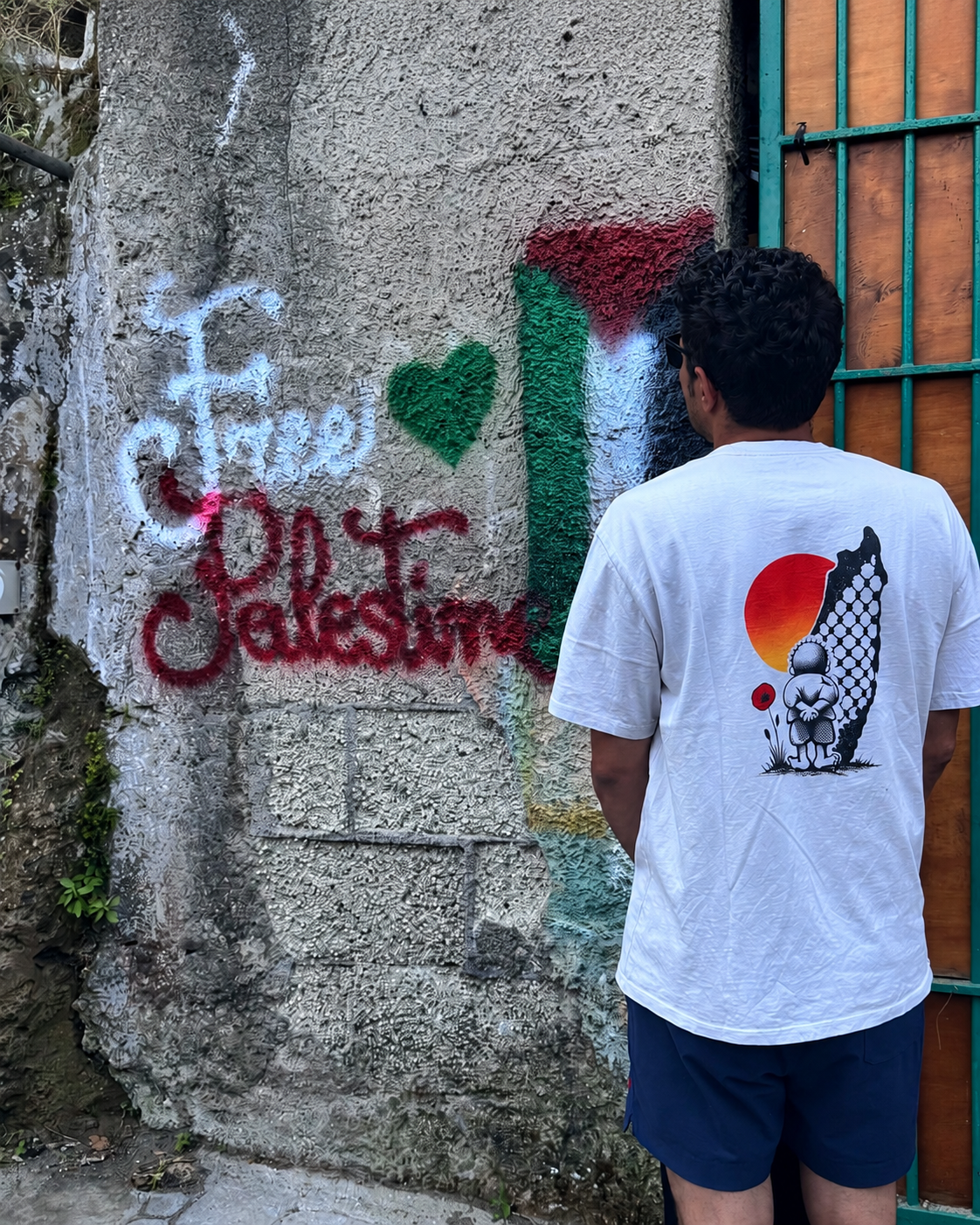 Man wearing the heavyweight Handala Palestinian T-shirt, shown from the back beside a wall with Free Palestine graffiti.