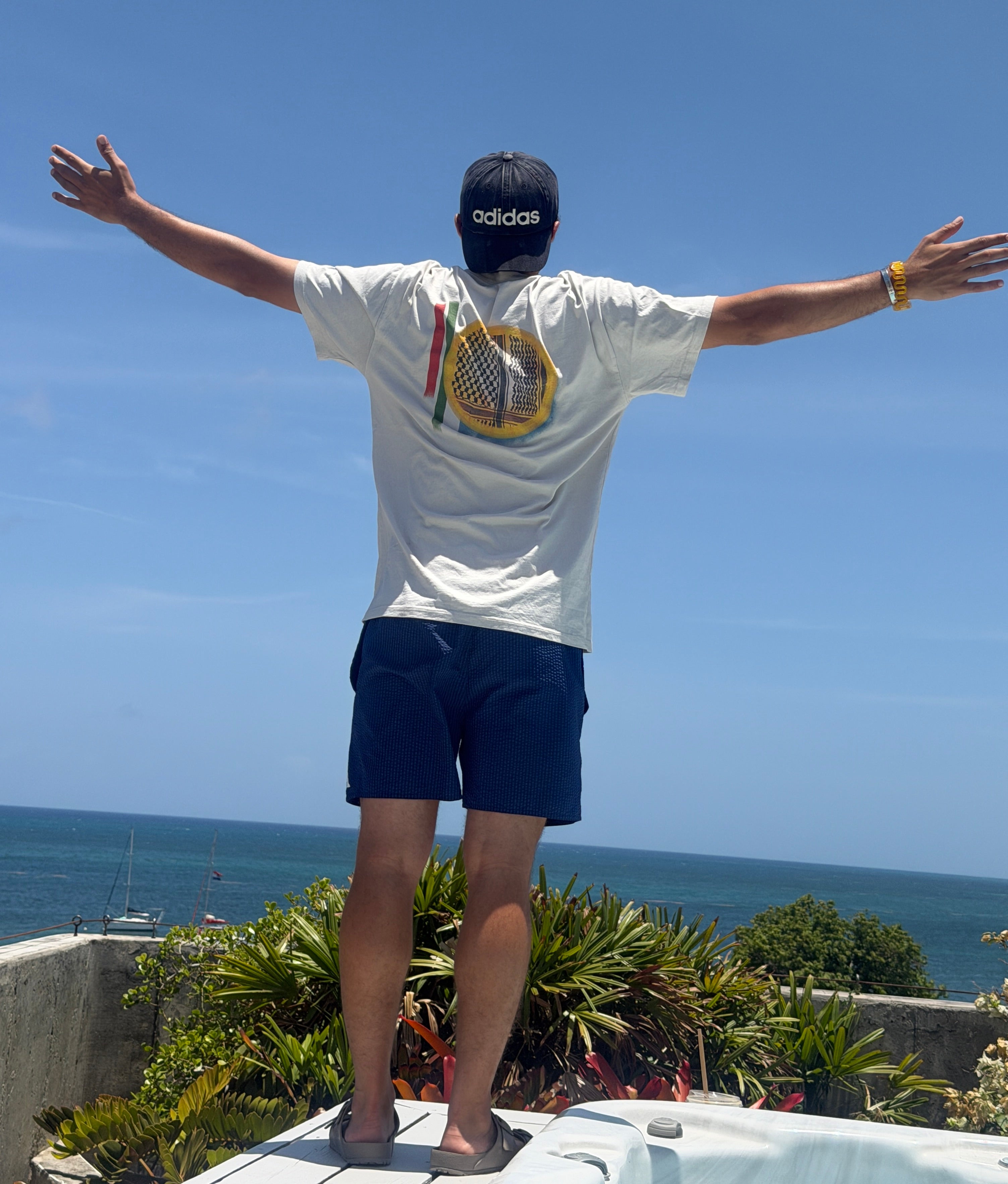 Man wearing the cream heavyweight Kufiyyeh Sun Palestinian T-shirt, shown from the back with arms outstretched by the ocean.