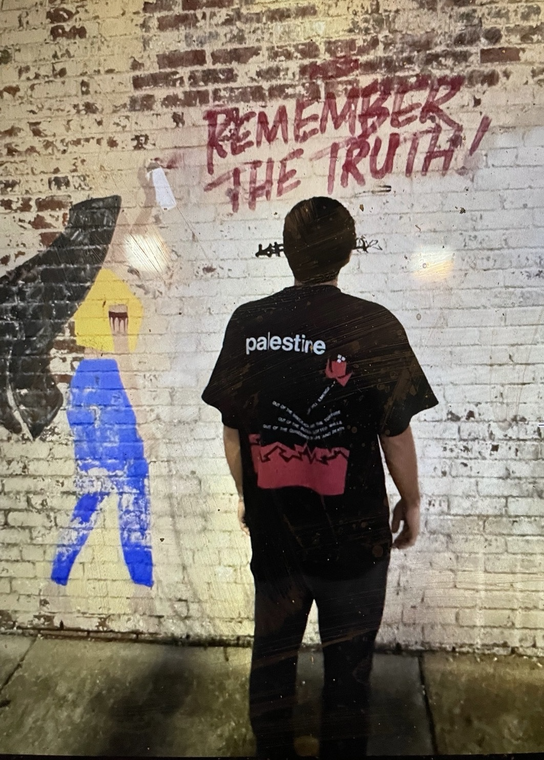 Man wearing the black heavyweight Life Will Emerge Palestinian T-shirt, shown from the back in front of a graffiti-covered wall.