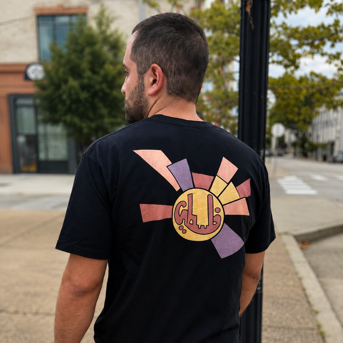 Man wearing black Sun Emblem Palestinian T-shirt, shown from the back on a city street.